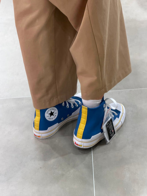 A person wearing blue, yellow, and white Converse high-top sneakers with tan pants, standing on a light gray tiled floor.