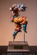 A detailed collectible statue of Goku in a dynamic martial arts pose, mounted on a wooden display base. The figure features his signature spiky silver hair, an orange gi, and blue wrist and boot accents, set against a solid warm brown background.