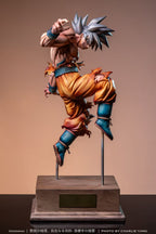 A detailed collectible statue of Goku in a dynamic martial arts pose, mounted on a wooden display base. The figure features his signature spiky silver hair, an orange gi, and blue wrist and boot accents, set against a solid warm brown background.