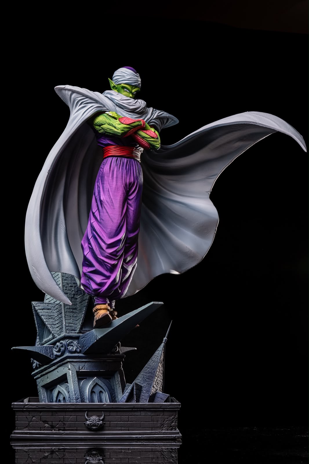 A detailed collectible statue of Piccolo from Dragon Ball Z, standing on a dark gothic pedestal with his cape dramatically spread, set against a solid black background.