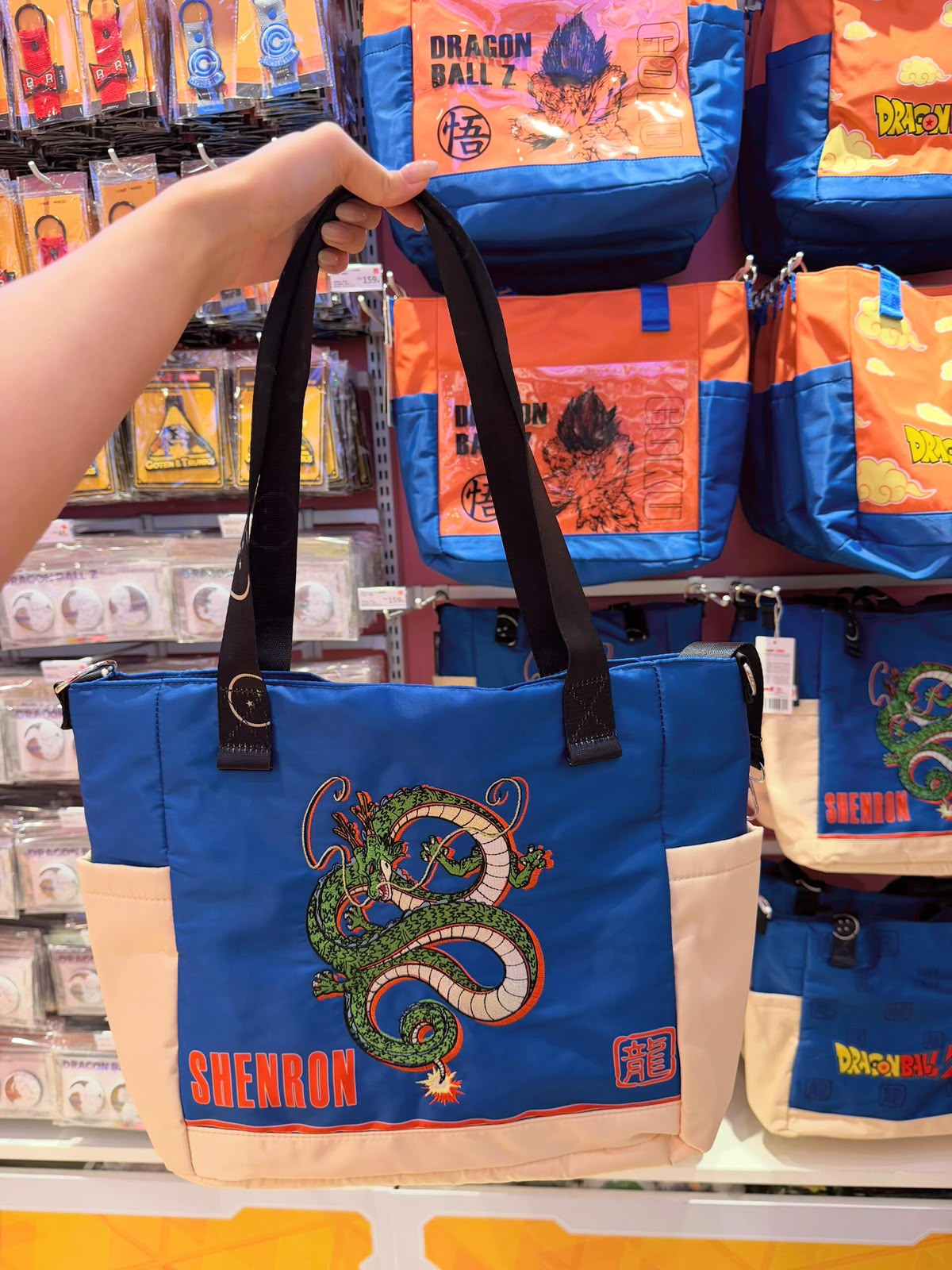 A person's hand holds up a blue and beige Dragon Ball Z tote bag featuring Shenron embroidered on the front, with other Dragon Ball Z bags displayed on the store shelves behind it.