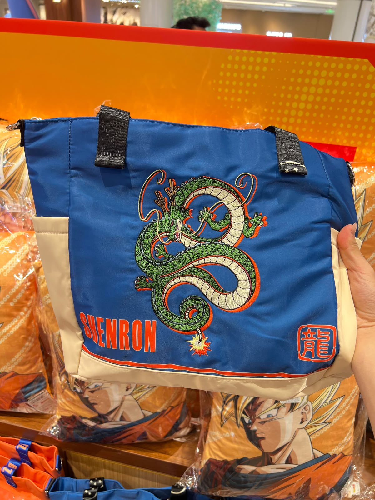 A blue Dragon Ball tote bag with an embroidered green and white dragon design, red 'GABENRON' text, and a red character emblem, held by a hand in a retail store setting.