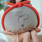 A person holding a large gray plush cushion shaped like a cat face with a red looped border and black embroidered facial features. The photo is taken indoors on a light sofa, with the person's hands and legs visible.