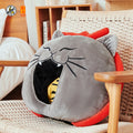 A round gray cat-shaped pet bed or cushion with a red outer ring, black whisker details, and an open mouth showing a yellow-and-black striped interior, placed on a woven chair in a bedroom setting.
