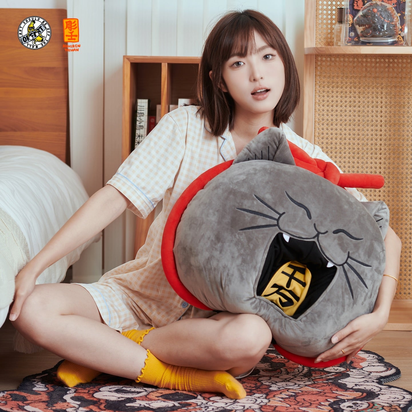 A woman sitting cross-legged on a patterned rug in a bedroom, holding a large plush cat-shaped pillow. The pillow is gray with a red collar, black whiskers and mouth details, and a yellow mouth interior containing a black Japanese character.