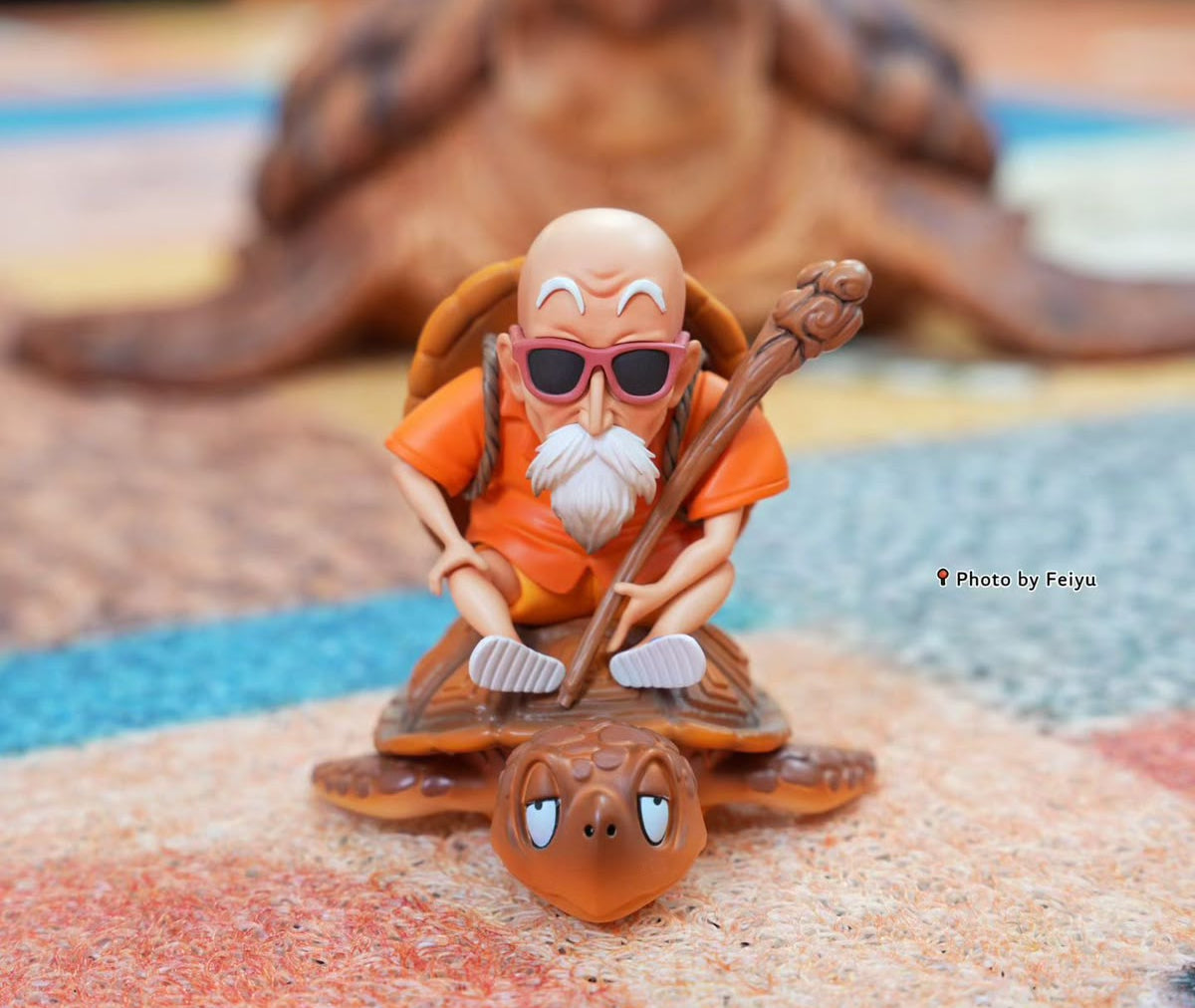 A small figurine of a bald man with a white beard wearing orange clothing and sunglasses, riding a brown turtle while holding a staff, displayed on a textured surface with a blurred background.